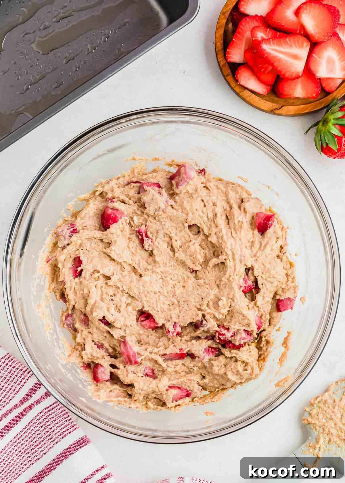 A glass bowl filled with strawberry bread batter. Nearby are a metal baking pan, a wooden bowl with sliced strawberries, and a red-striped kitchen towel on a light-colored countertop.