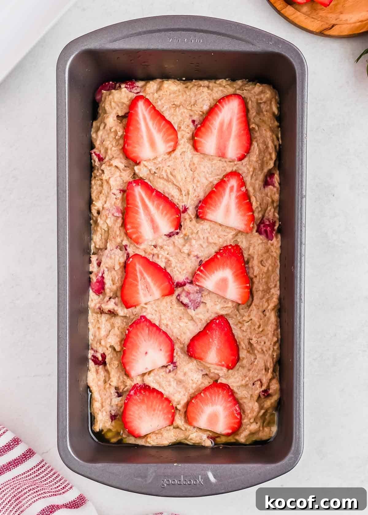 Loaf pan filled with unbaked strawberry bread batter, topped with sliced strawberries in a pattern on top. The pan is placed on a light surface, and a striped cloth is partially visible in the bottom left corner.