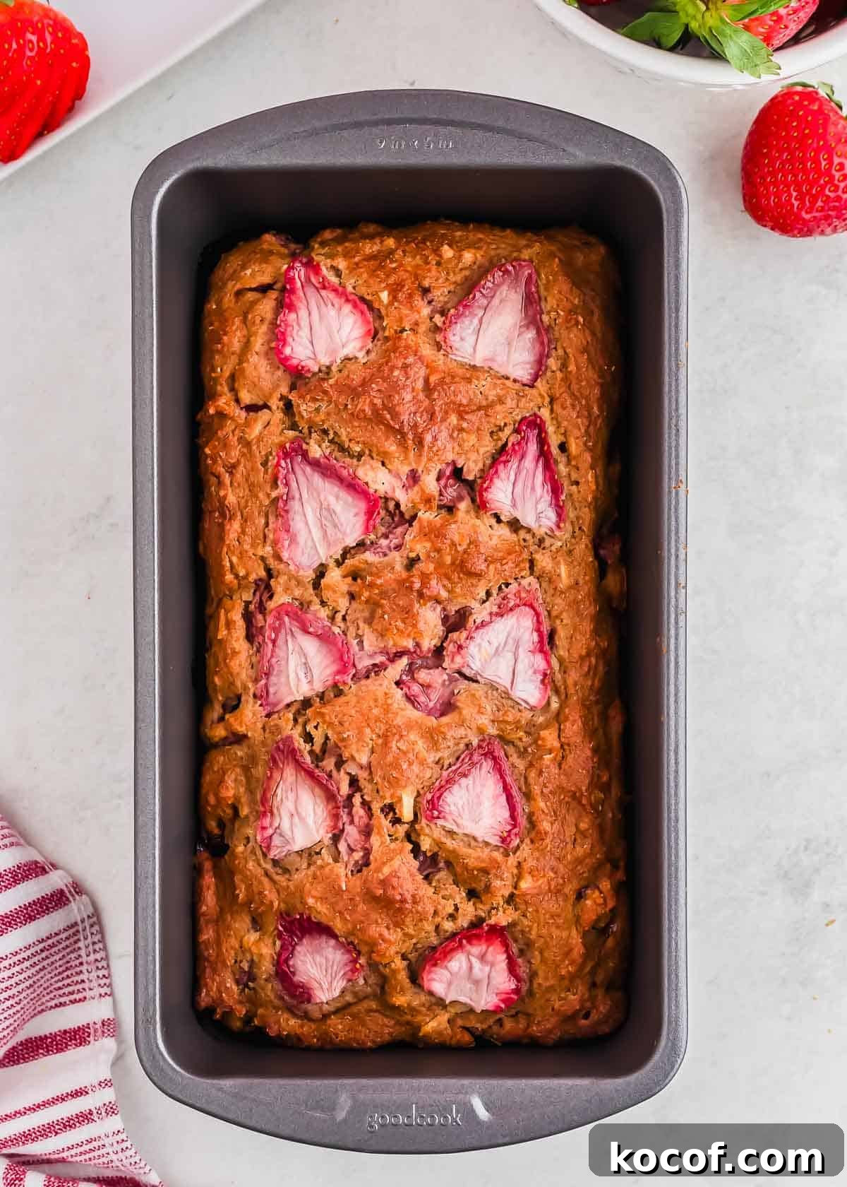 A loaf of strawberry bread in a rectangular baking pan. The bread is golden brown with visible slices of strawberries on top. A red and white striped cloth and fresh strawberries are nearby on a light surface.