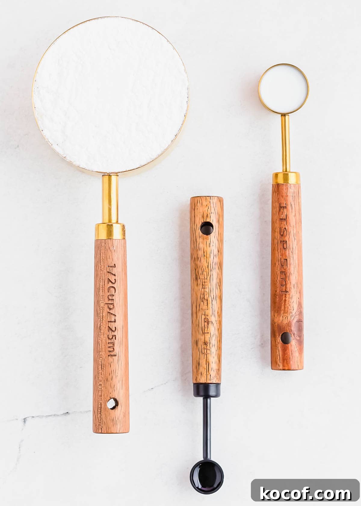 Three measuring spoons with wooden handles are arranged on a white surface. The largest contains powdered sugar, while the other two contain milk and vanilla to make a glaze. Each spoon handle has a hole for hanging and measurement markings etched into the wood.