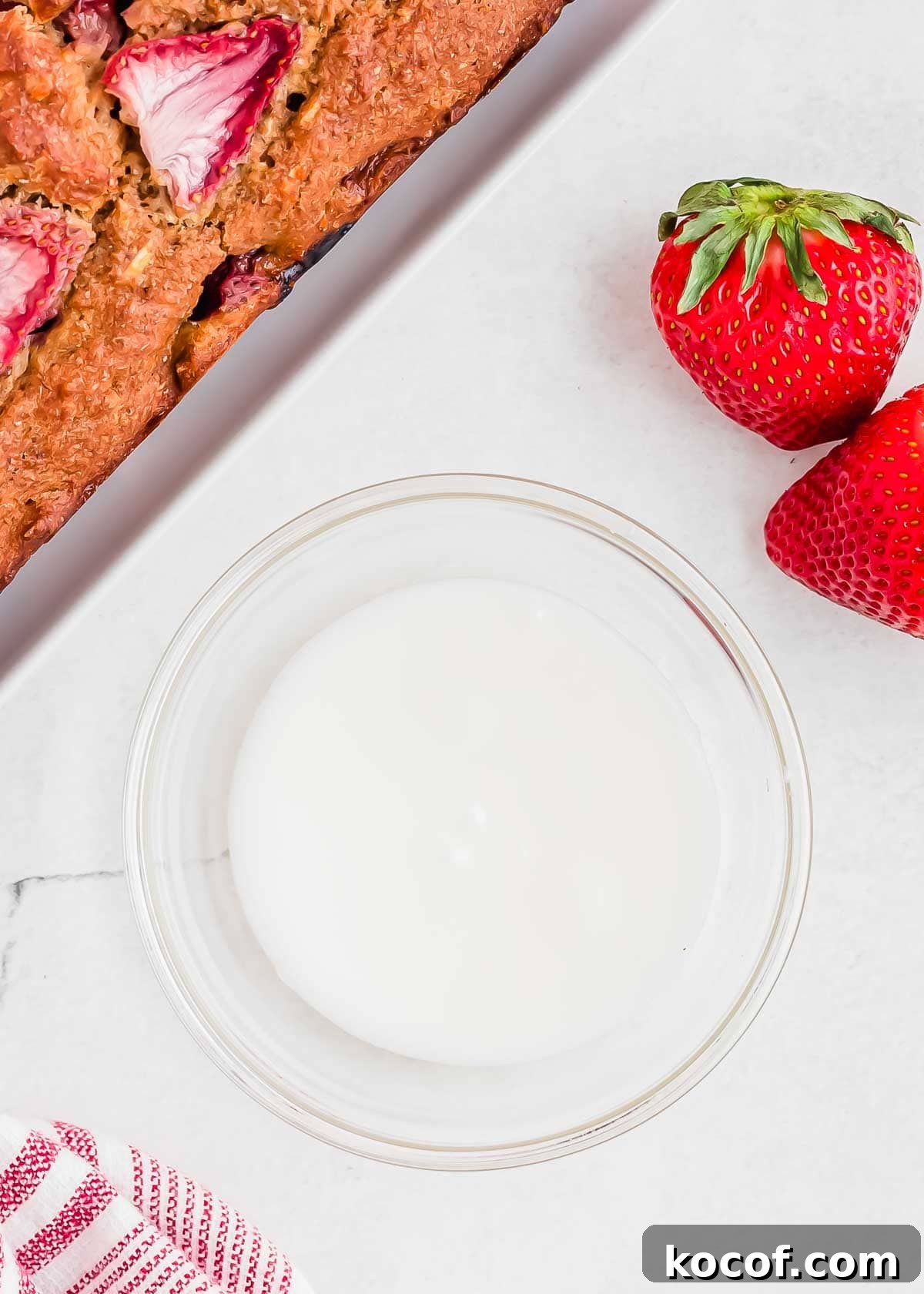 A glass bowl filled with glaze is on a white surface. To the side, there are two fresh strawberries with green leaves. Part of a baked strawberry bread is visible in the top left corner. A striped cloth is partially seen in the bottom left.
