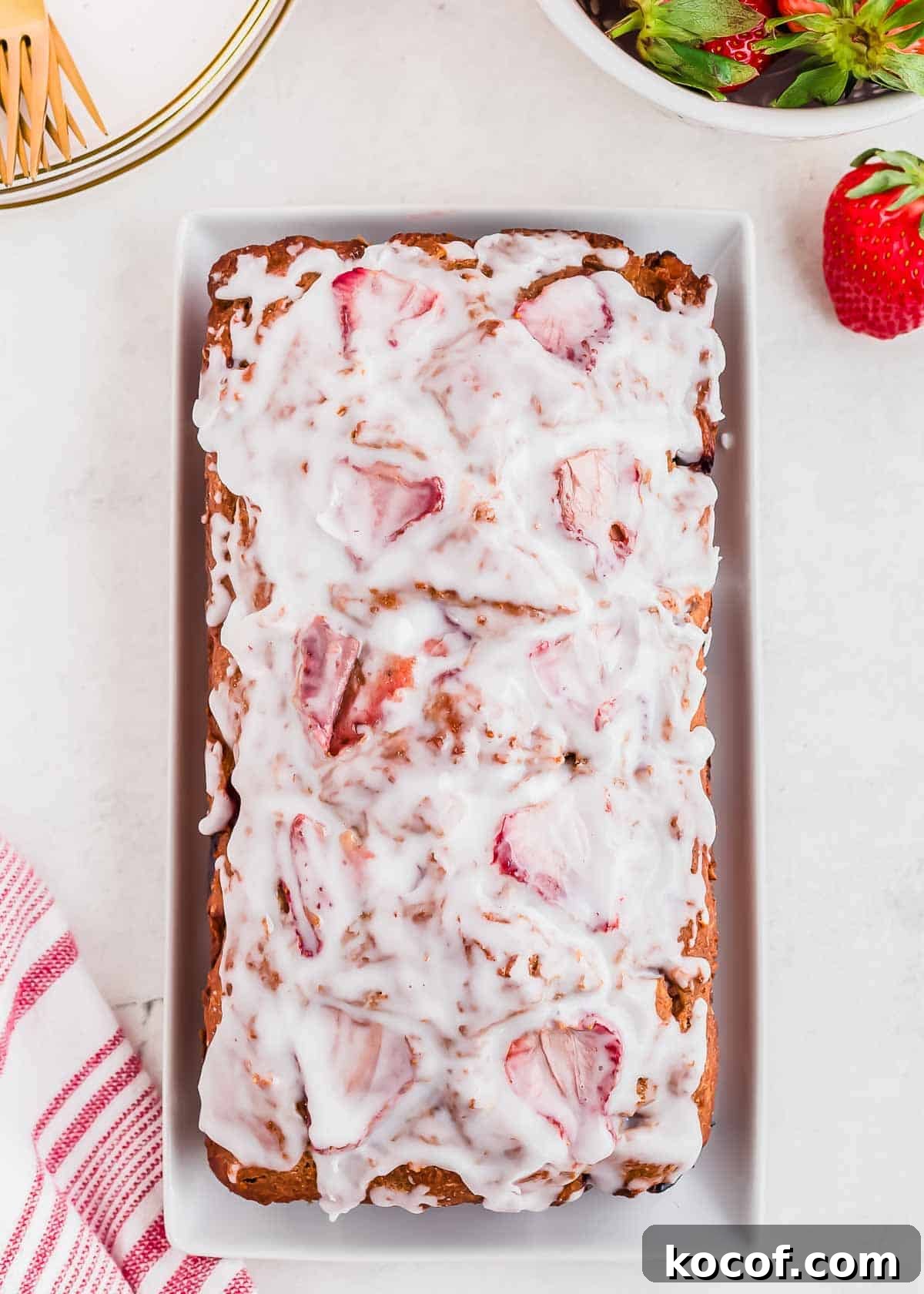 A glazed strawberry bread loaf on a rectangular white plate. The bread is topped with a white glaze and visible strawberry slices. A bowl of whole strawberries and a red-striped cloth are nearby on the light-colored surface.