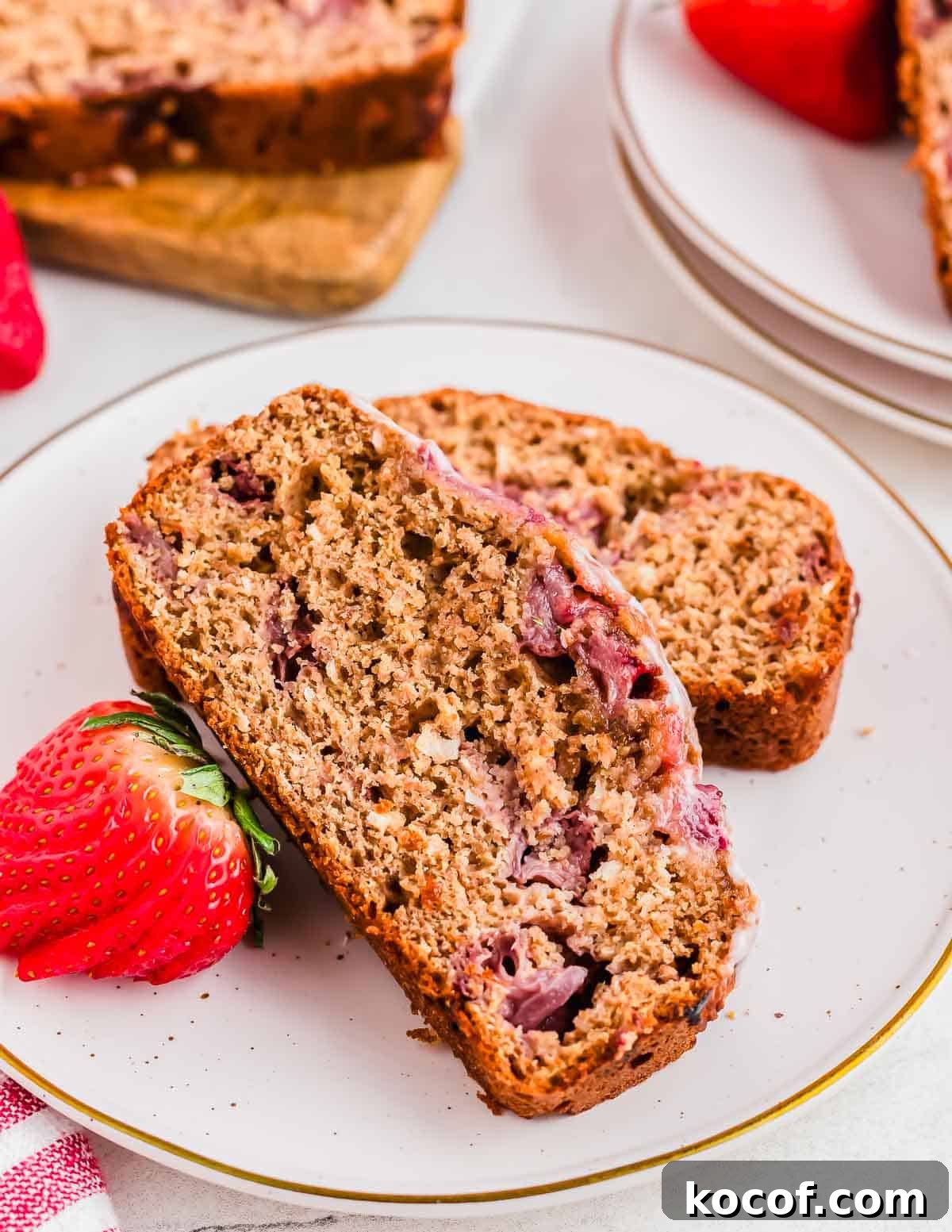 Two slices of strawberry bread are placed on a white plate, accompanied by a fresh strawberry. The bread features visible chunks of strawberries and a moist, dense texture. Another plate and a cutting board with more bread are in the background.