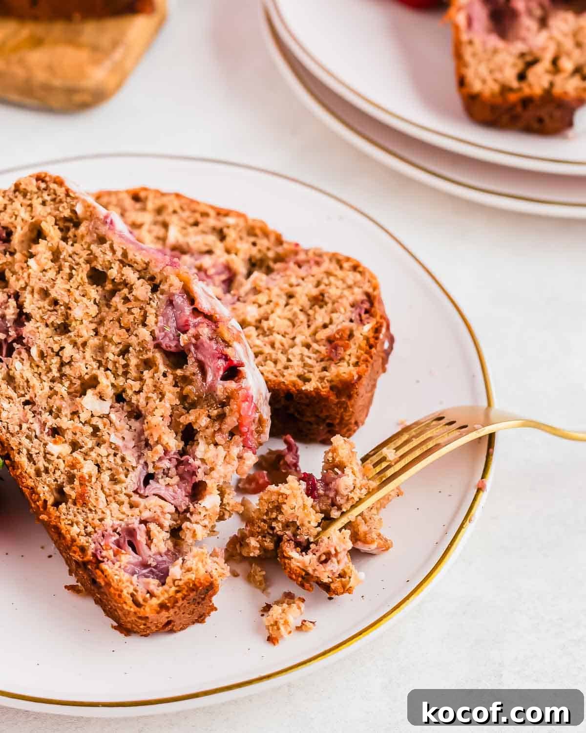 Slices of a crumbly moist strawberry bread with visible pieces of fruit, are placed on a white plate with a gold trim. A fork rests on the plate, breaking off a piece of the bread. The background includes another plate and blurred elements.