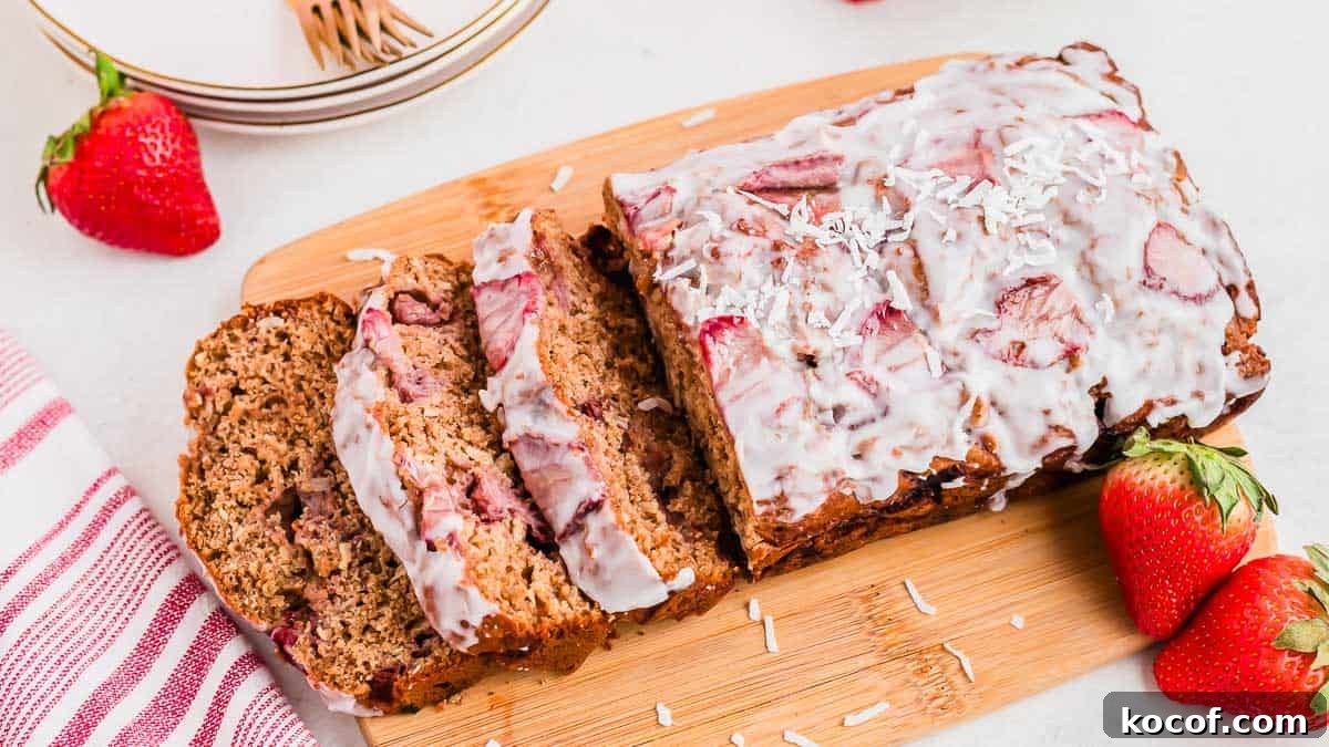 A sliced loaf of strawberry bread with icing on a wooden cutting board. Surrounded by fresh strawberries and a red-striped cloth. Some shredded coconut sprinkled on top adds texture to the glaze. White plates and gold forks are in the background.
