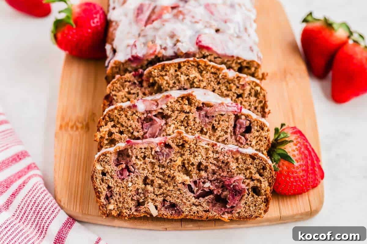 Sliced strawberry bread with glaze on a wooden cutting board, garnished with fresh strawberries. A red and white striped cloth napkin is visible on the side.