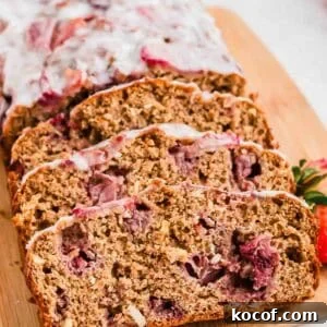 Sliced loaf of strawberry bread with a white glaze on top, resting on a wooden cutting board. The bread is visibly dense, with chunks of strawberries throughout. A few fresh strawberries are placed beside the loaf for garnish.