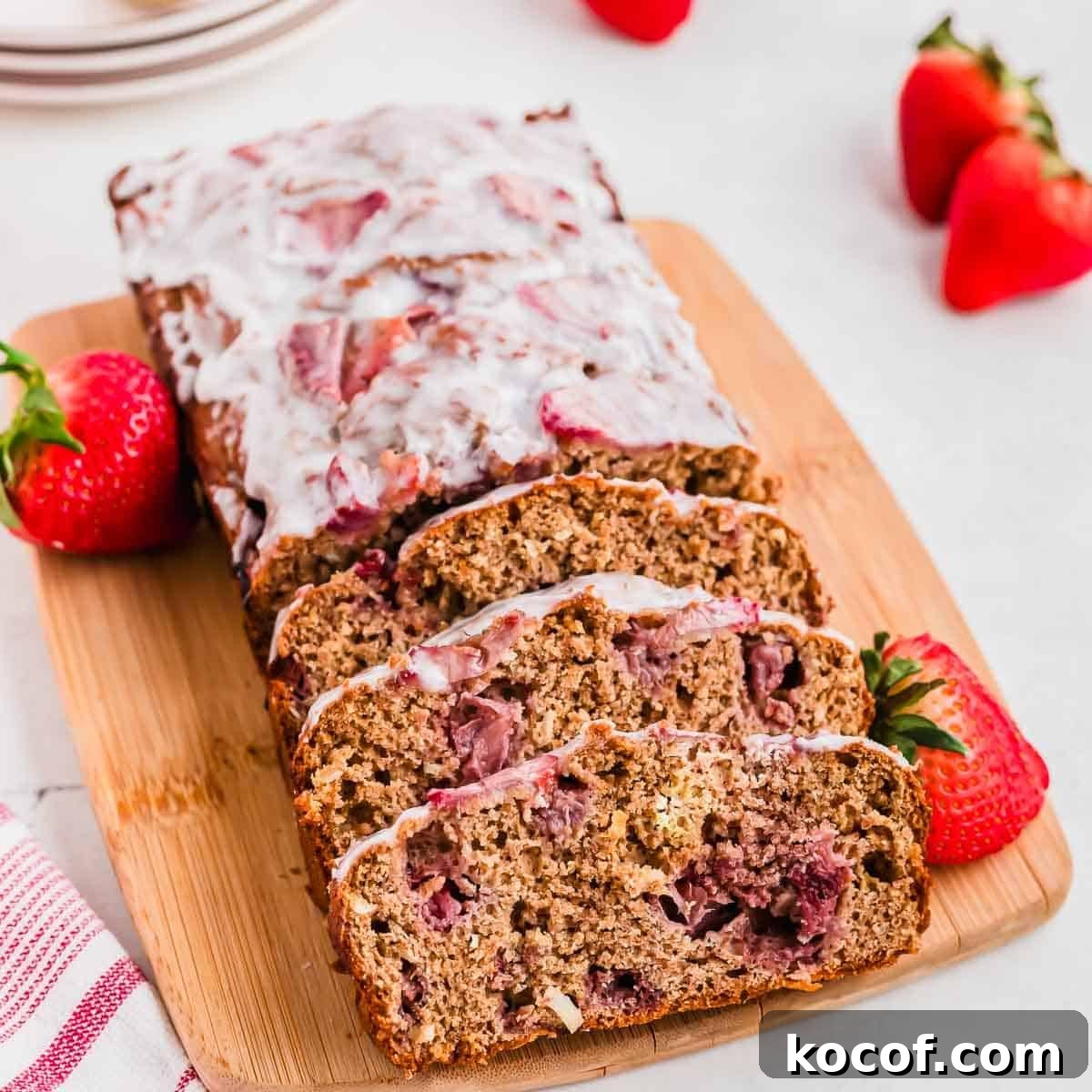 A sliced loaf of strawberry bread with glaze on a wooden board, surrounded by whole strawberries. A red and white striped cloth is in the foreground, and a stack of plates is partially visible in the background.