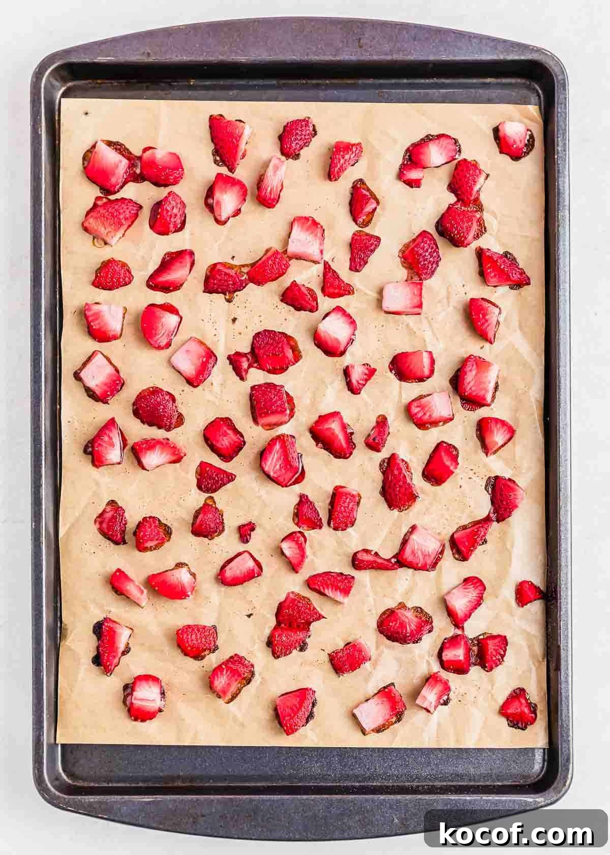 Chunks of strawberries are spread evenly on a parchment-lined baking sheet, after roasting. The vibrant red pieces are scattered randomly, contrasting with the light brown sheet.
