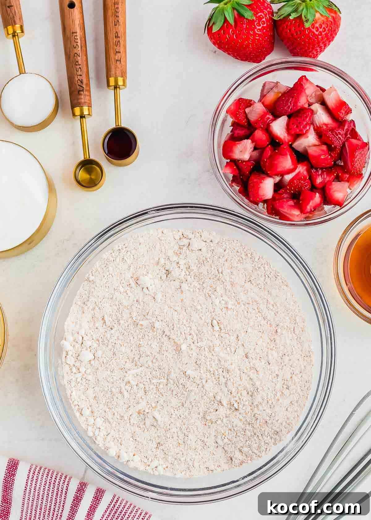 Top view of baking ingredients, including a bowl of flour, a bowl of chopped strawberries, measuring spoons, a cup of sugar, a cup of brown liquid, and whole strawberries. A striped cloth is partially visible at the bottom.