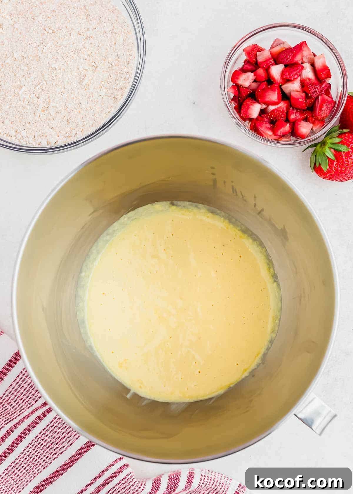 A mixing bowl with a yellow bread batter is placed on a countertop. Next to it, there are small bowls containing chopped strawberries and a dry ingredient mixture. A red and white striped cloth is partially visible in the corner.