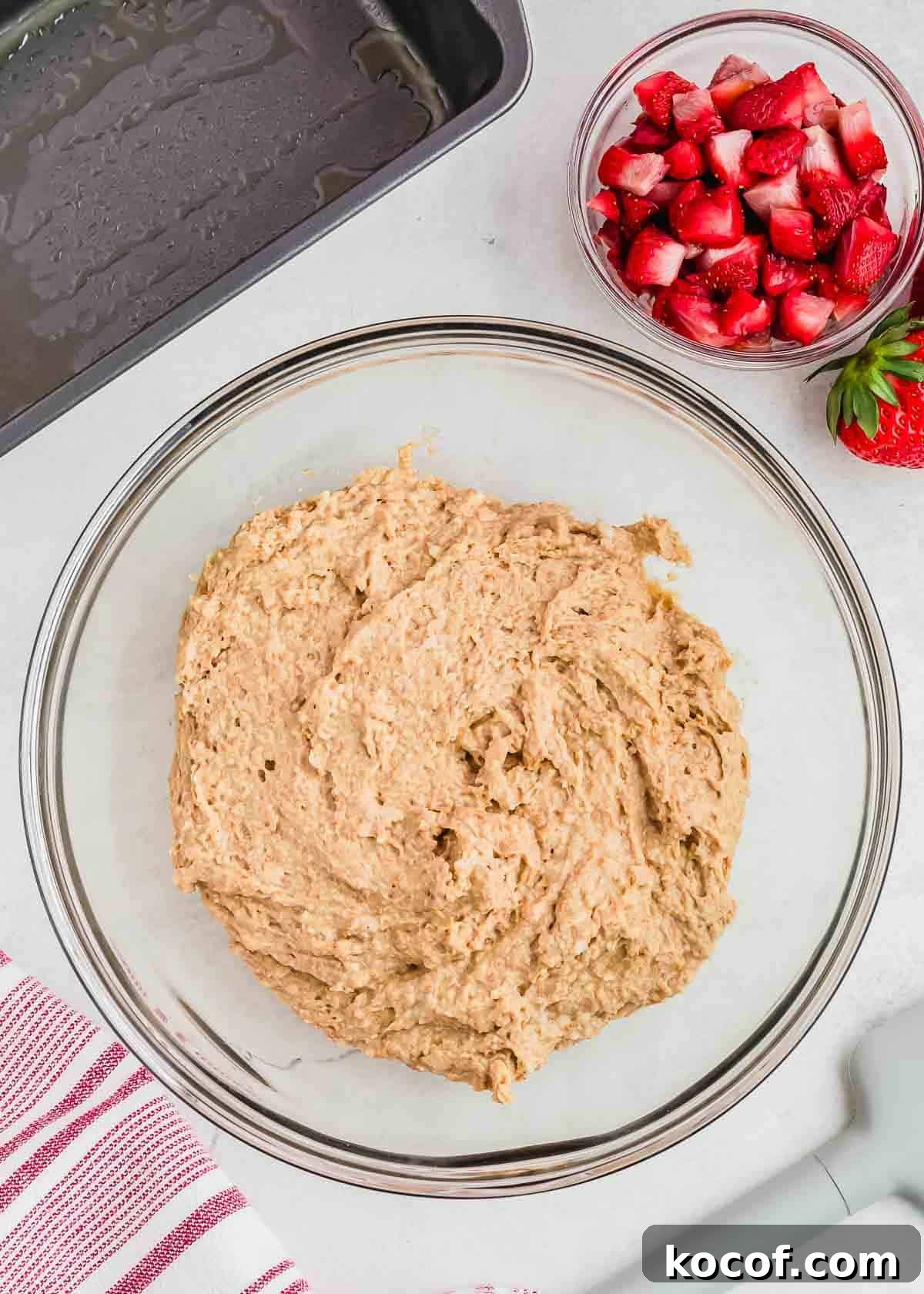 A glass bowl filled with light brown bread batter sits on a light gray surface. Nearby, a smaller bowl contains chopped strawberries. A striped red and white cloth and a gray baking dish are also visible.