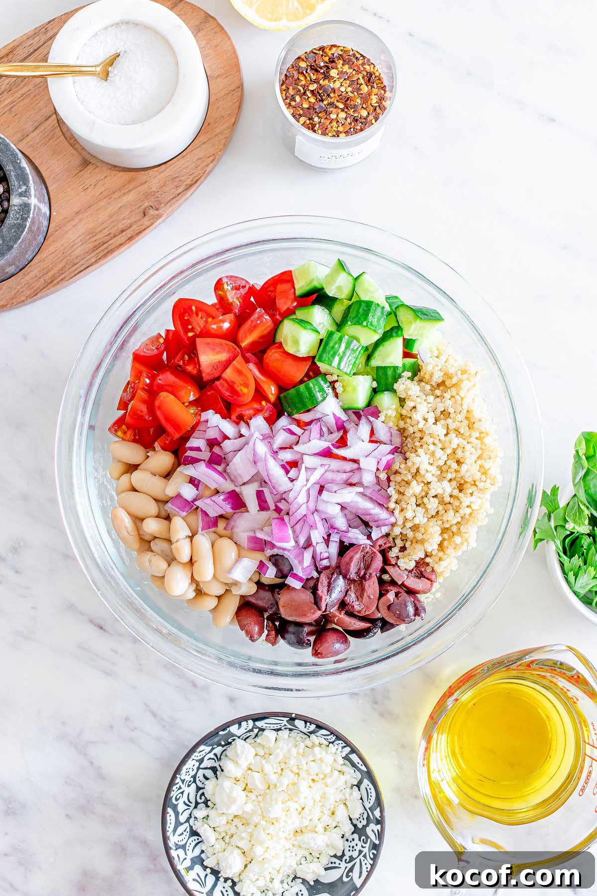 Mediterranean Quinoa Salad with Feta, Cucumbers & Olives 14 A glass bowl filled with chopped tomatoes, cucumber, onion, beans, olives, and quinoa creates a vibrant Mediterranean Quinoa Salad on a marble counter.