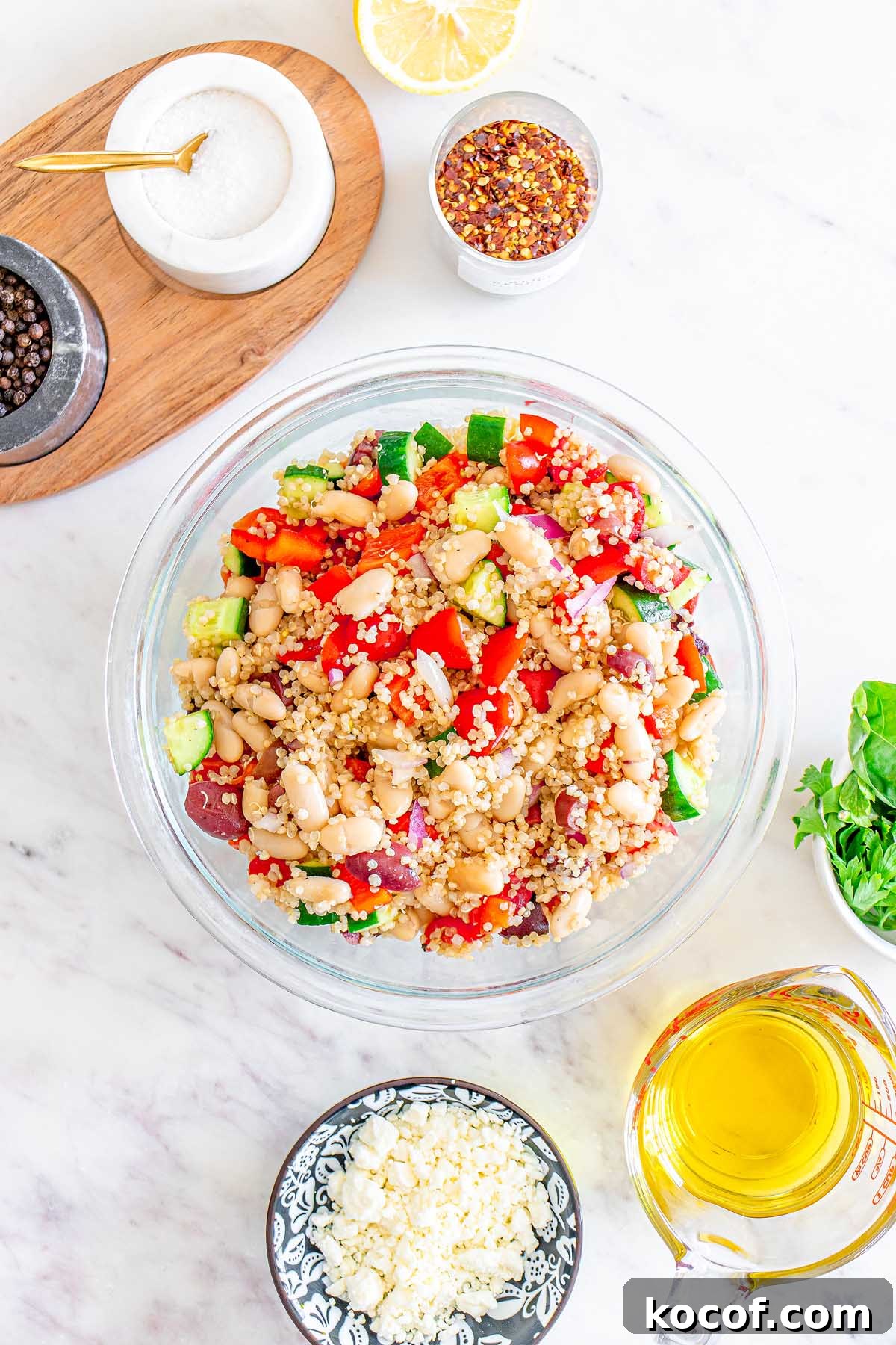 Mediterranean Quinoa Salad with Feta, Cucumbers & Olives 15 A glass bowl of Mediterranean Quinoa Salad with beans, tomatoes, cucumber, and onions sits surrounded by fresh ingredients on a white table.