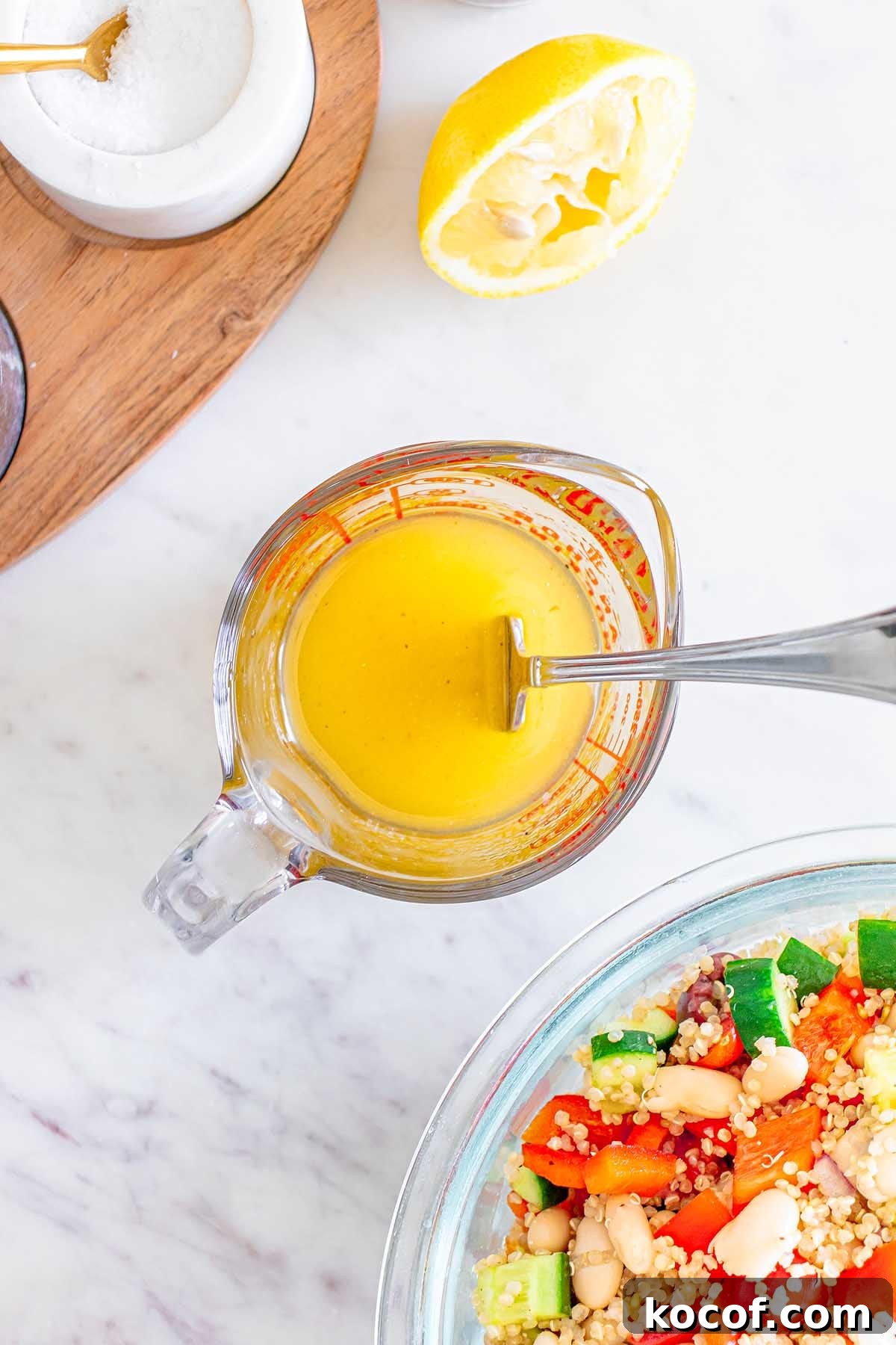 Mediterranean Quinoa Salad with Feta, Cucumbers & Olives 16 Glass measuring cup with yellow dressing and fork, next to a bowl of Mediterranean Quinoa Salad and a squeezed lemon on a marble counter.