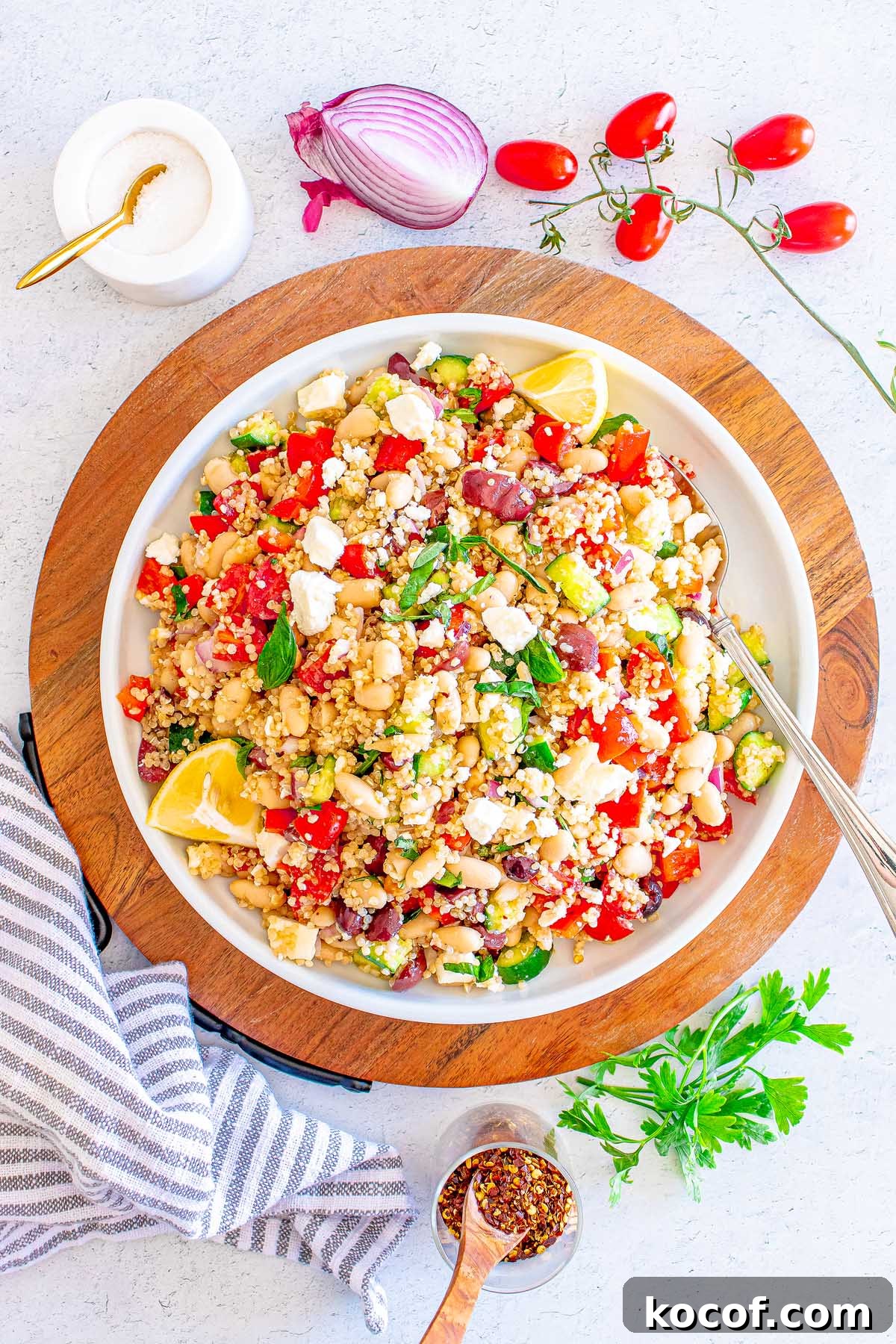 Mediterranean Quinoa Salad with Feta, Cucumbers & Olives 6 A bowl of Mediterranean Quinoa Salad with beans, feta, tomatoes, and lemon wedges sits on a wooden board.