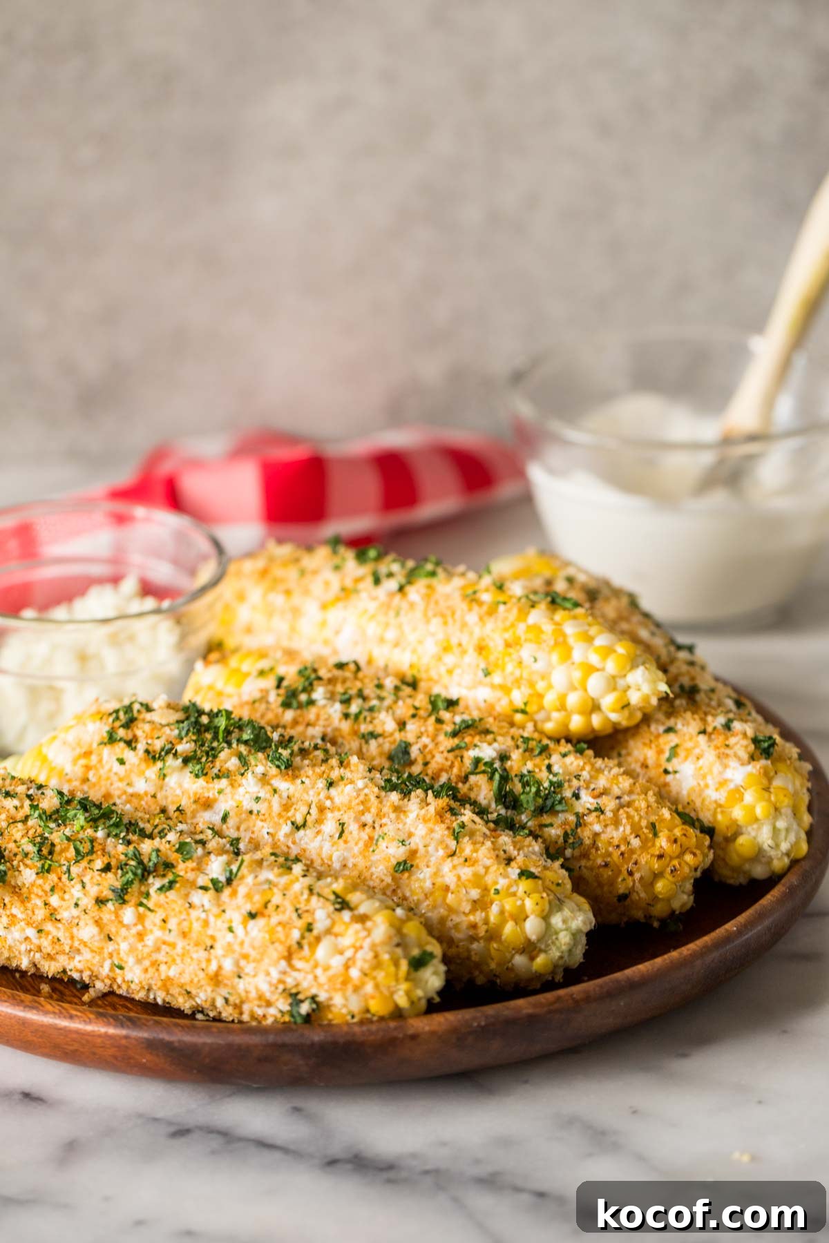 Mexican Street Corn With Panko and a small glass dish of cotija cheese on a wood platter, with a glass bowl of the mayonnaise mixture in the background.