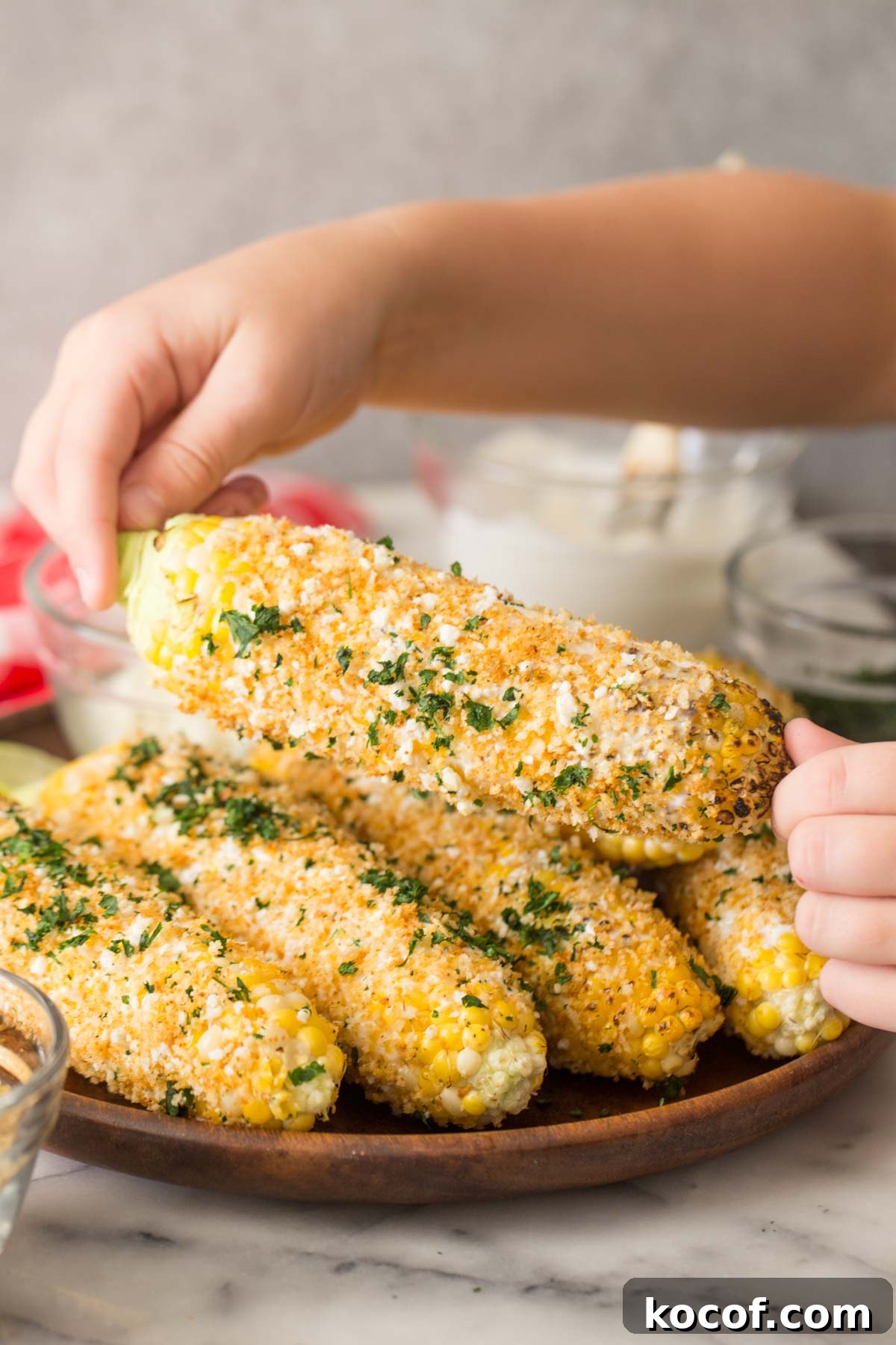 Two hands holding an ear of Mexican Street Corn With Panko, with more ears of corn on a wood platter in the background.