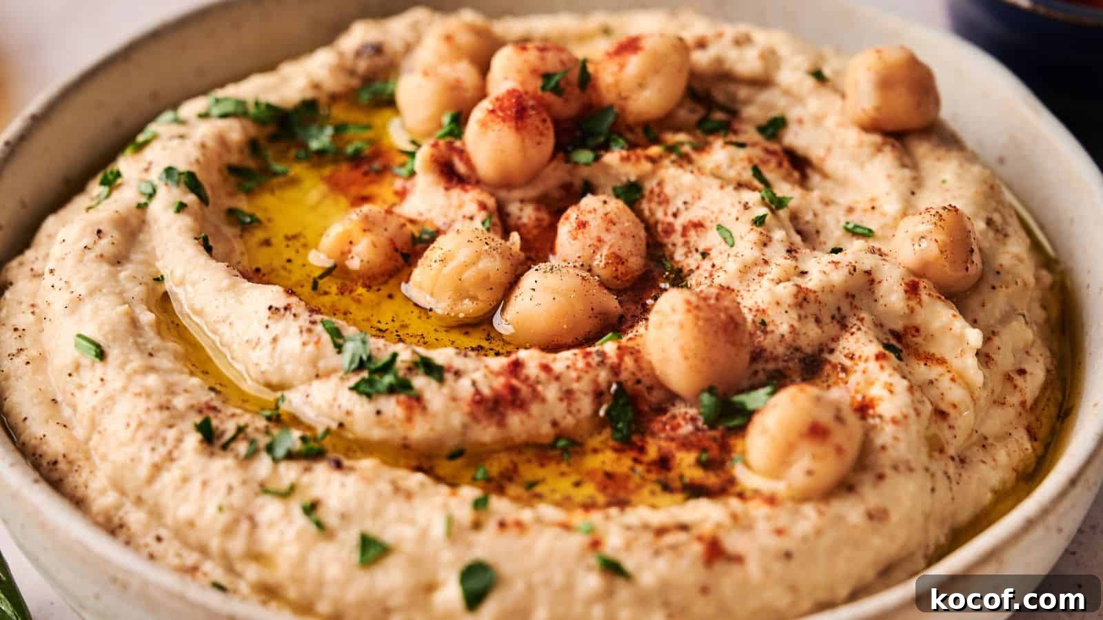 A bowl of hummus garnished with whole chickpeas, chopped parsley, paprika, and a drizzle of olive oil.