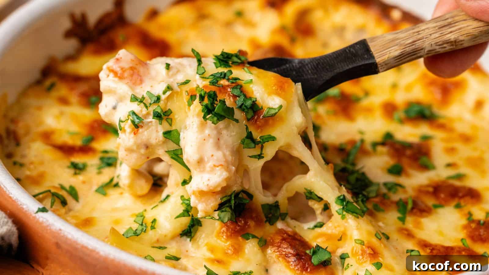 A spoon lifts a portion of cheesy baked pasta topped with chopped parsley from a casserole dish.