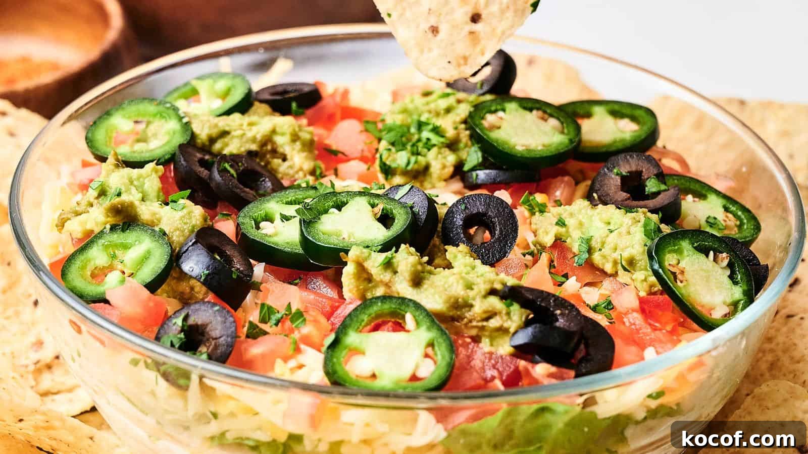 Glass bowl with layered dip topped with guacamole, sliced jalapeños, black olives, and tomatoes. A tortilla chip is dipping into the bowl.