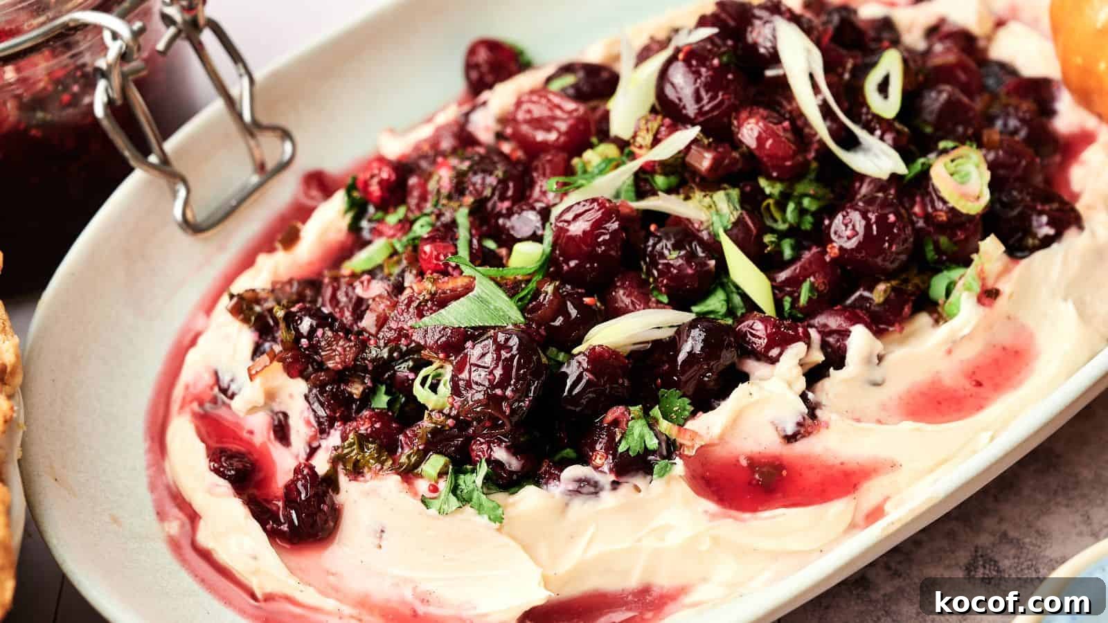 A platter of whipped cheese topped with roasted cranberries, chopped herbs, and sliced green onions.