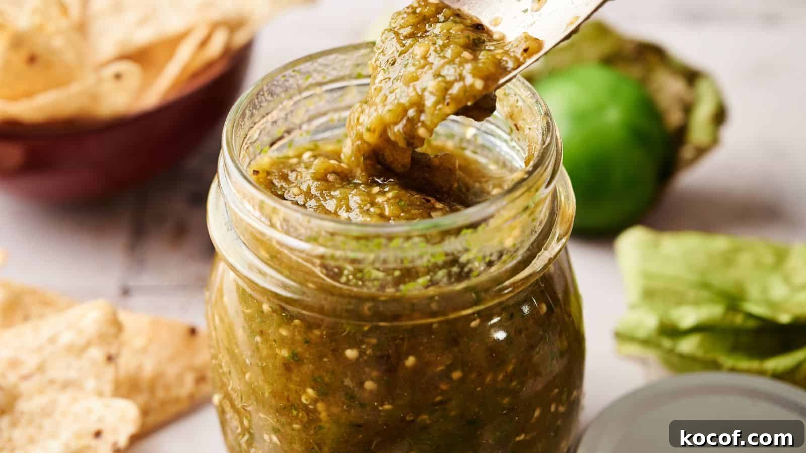 A jar of green salsa with a spoon lifting some out; tortilla chips and a tomatillo are visible in the background.