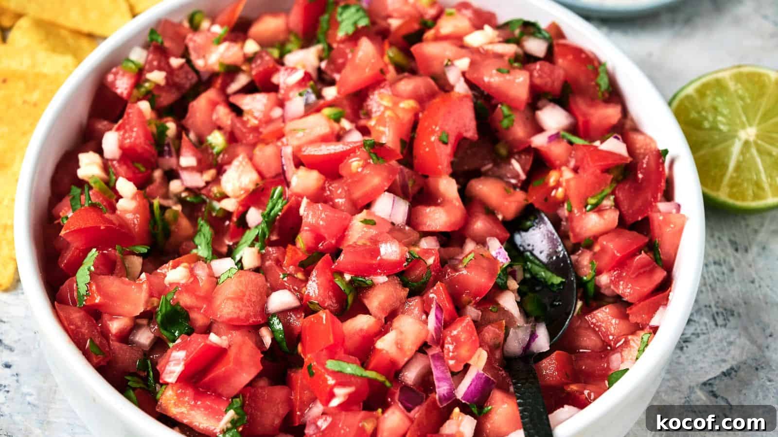 A bowl of pico de gallo with chopped tomatoes, onions, cilantro, and jalapeños with a spoon inside, next to a lime and tortilla chips.
