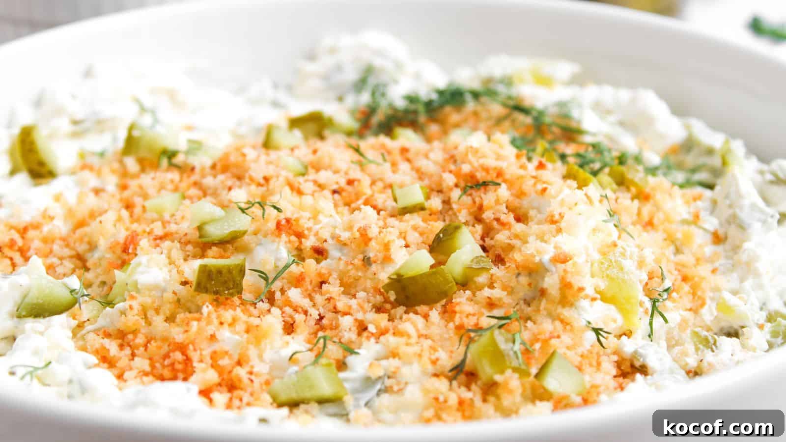 A close-up of a creamy dip topped with toasted breadcrumbs, chopped pickles, and fresh dill in a white bowl.