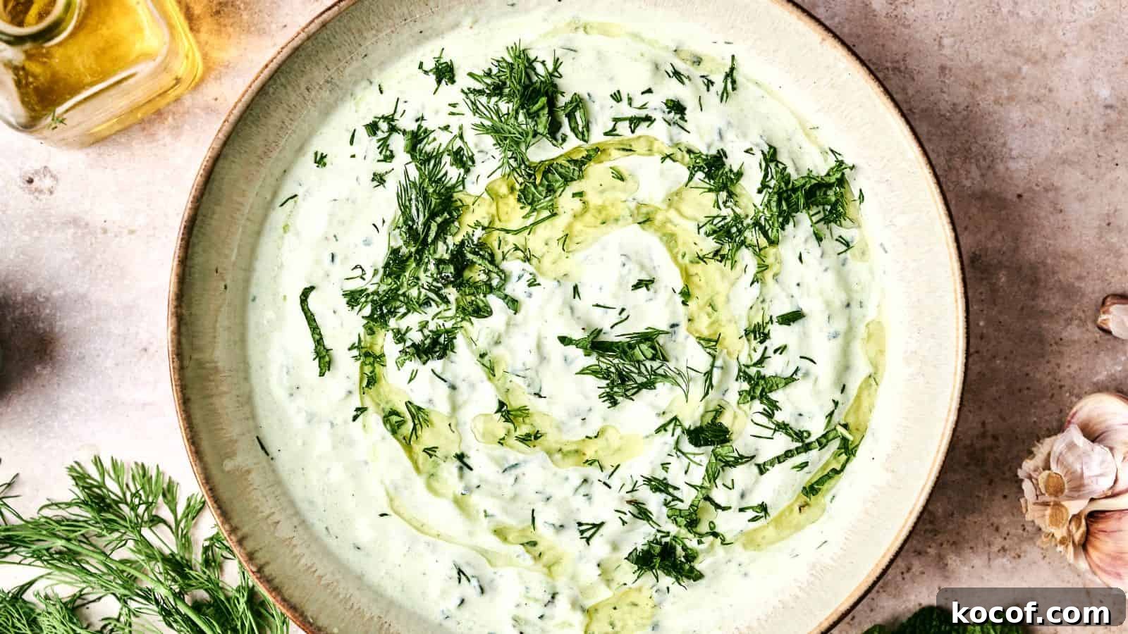 A bowl of creamy tzatziki sauce garnished with chopped dill and a swirl of olive oil. Fresh dill and garlic are nearby.