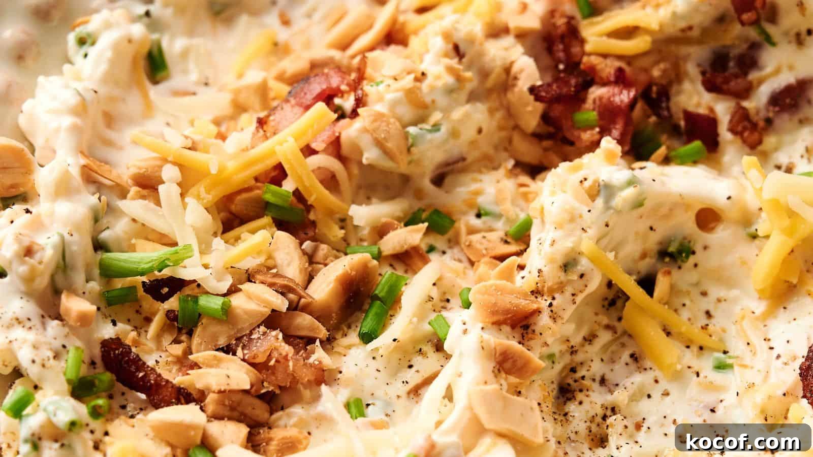 Close-up of a creamy dish topped with shredded cheese, chopped nuts, bacon bits, and sliced green onions.