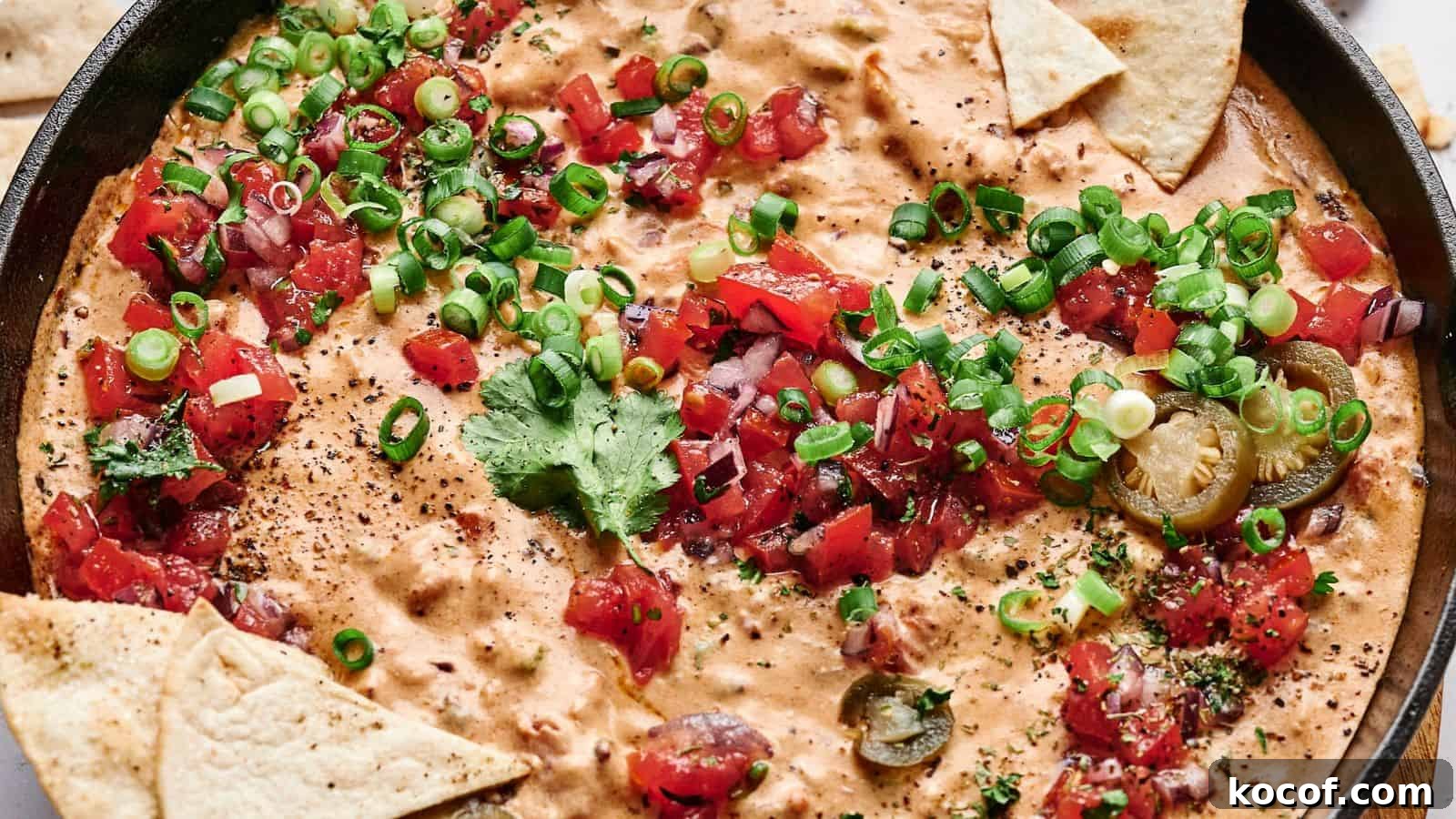 A skillet filled with creamy cheese dip topped with chopped tomatoes, green onions, cilantro, and sliced jalapeños, surrounded by triangular tortilla chips.
