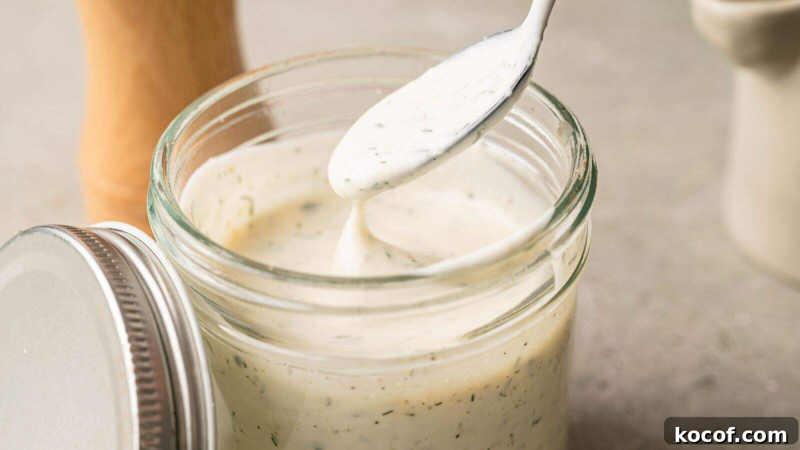 A spoon being dipped into a glass jar filled with creamy white dressing, with herbs visible throughout the mixture.