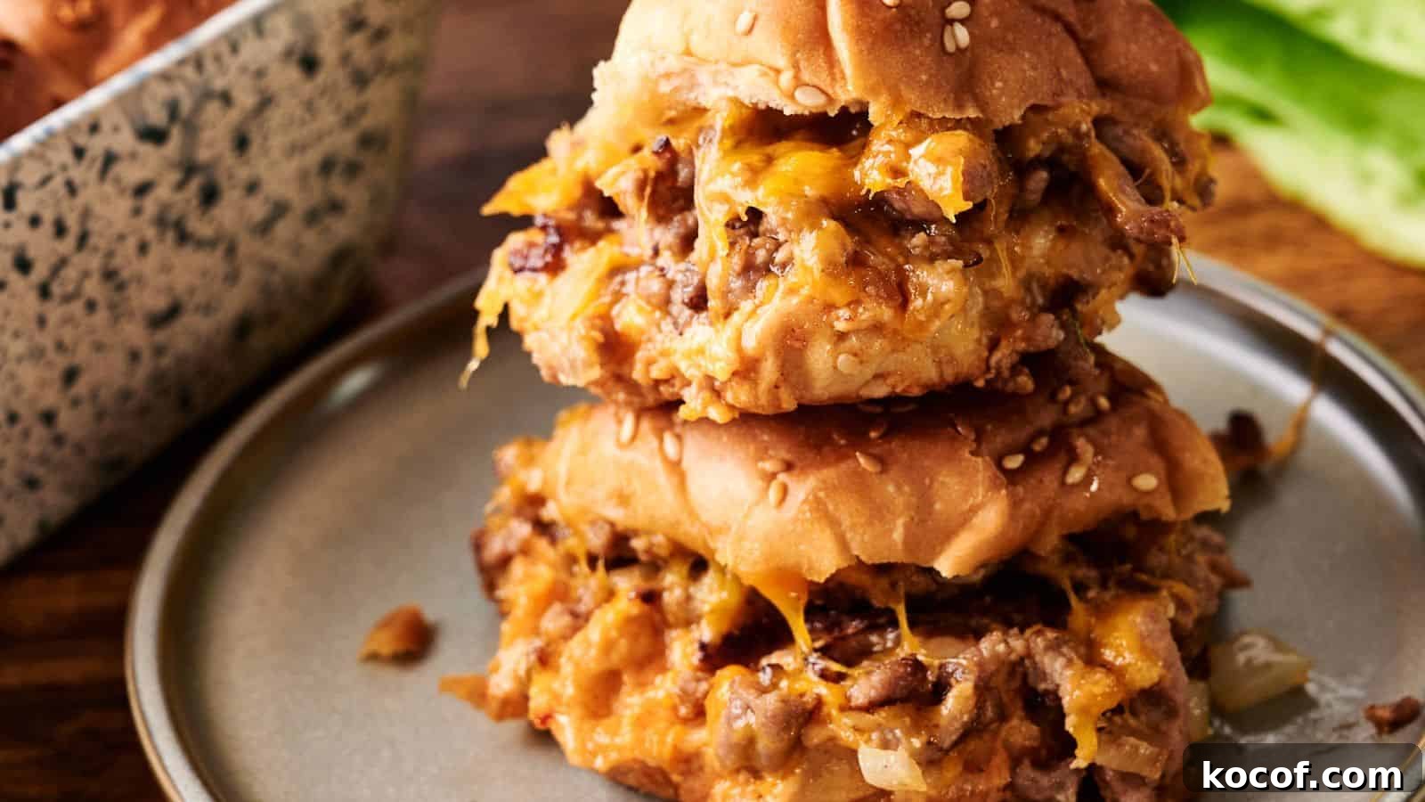 Two sloppy joe sandwiches with melted cheese on sesame seed buns are stacked on a gray plate.