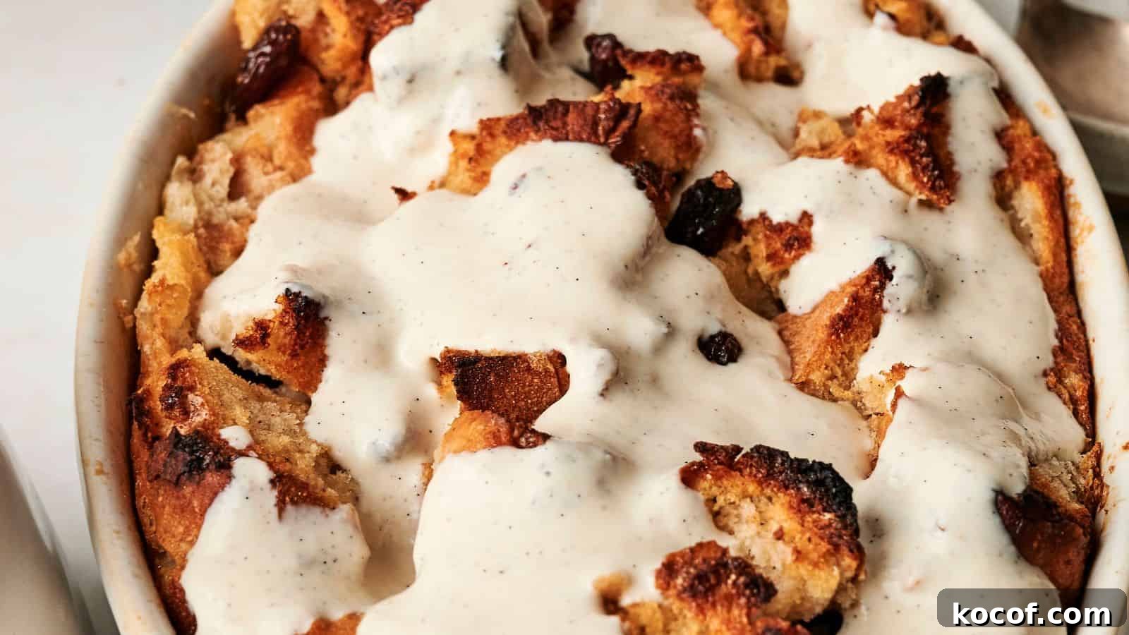 A close-up of bread pudding topped with creamy vanilla sauce in a white baking dish. The pudding features toasted bread pieces and visible raisins.
