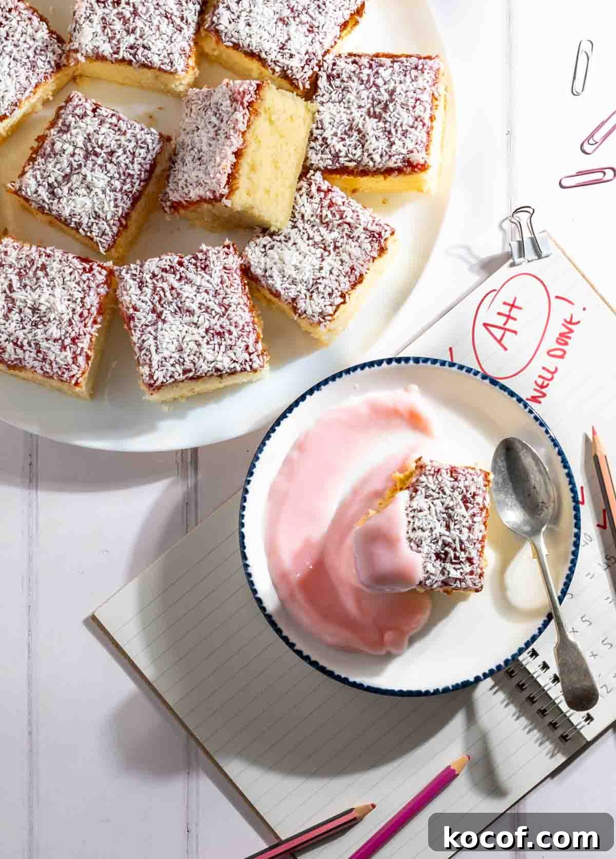 Squares of jam and coconut sponge on a white plate alongside a slice served with pink custard.