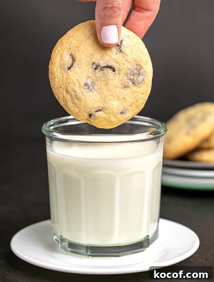 Fingers about to dip a chocolate chip cookie in a large glass of milk on a small white plate